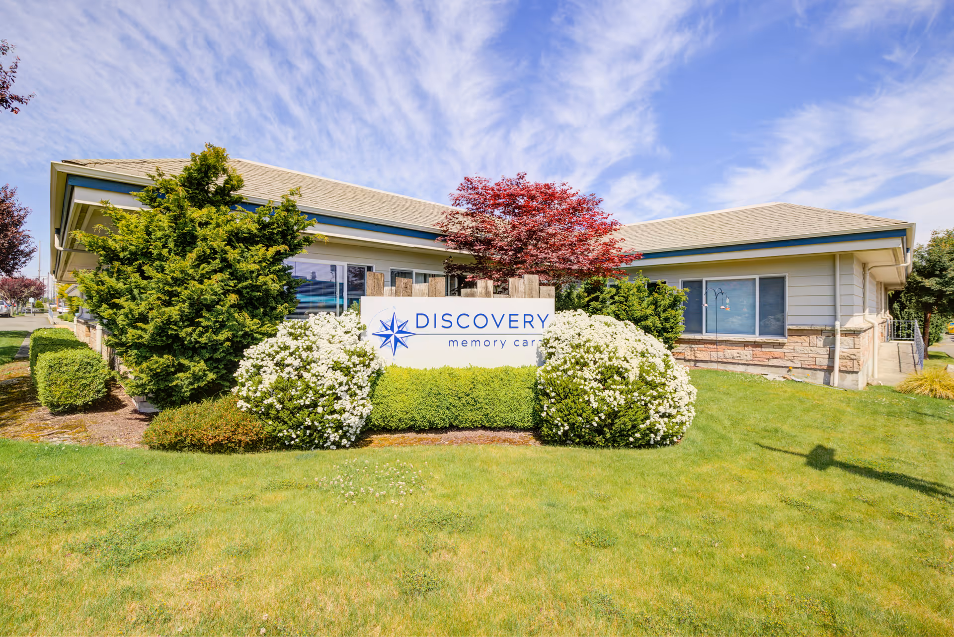 Front view of a single-story memory care building with a 'Discovery memory care' sign surrounded by shrubs and lawn under a blue sky.