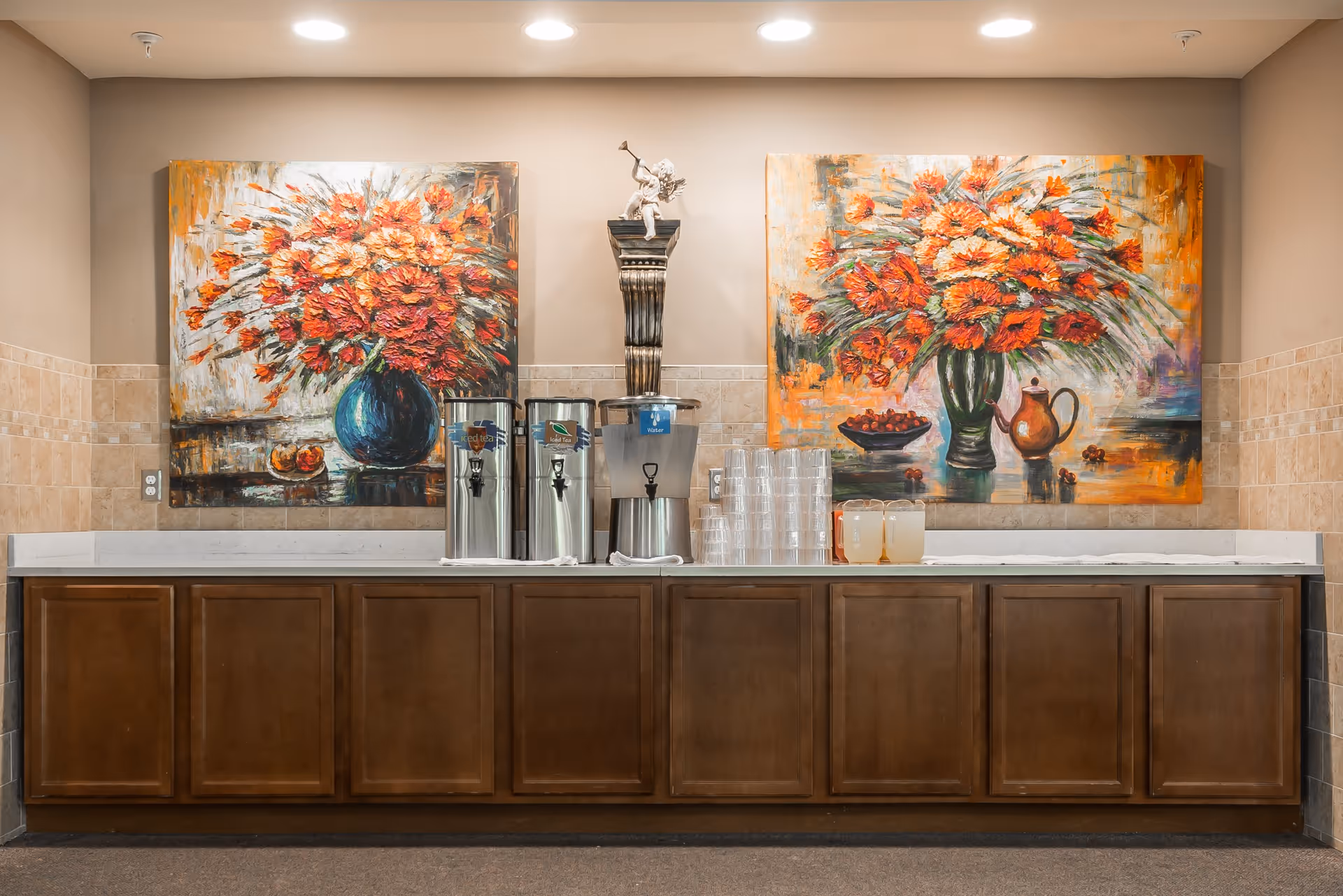 A beverage station with three large drink dispensers labeled iced tea, hot water, and water, along with stacks of clear plastic cups and two pitchers of lemonade on a long wooden counter. The wall behind features two large colorful paintings of flower arrangements in vases and a small statue of a cherub playing a trumpet on a pedestal between the paintings.