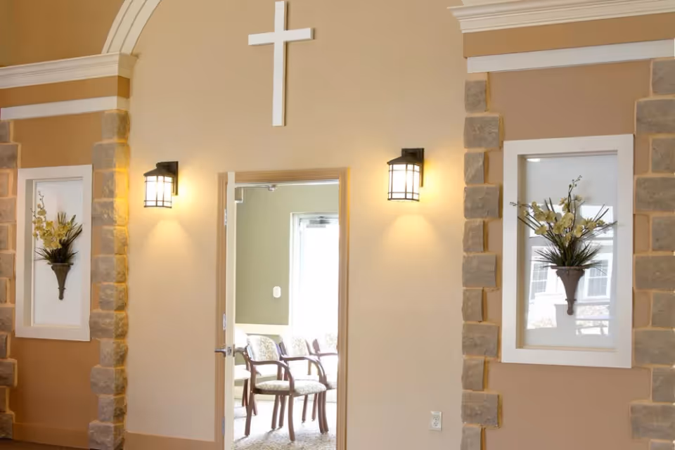 Interior view of a room with beige walls, stone accents around two recessed wall niches holding floral arrangements, two wall-mounted lantern-style lights, and a white cross mounted above an open doorway leading to a room with several chairs.
