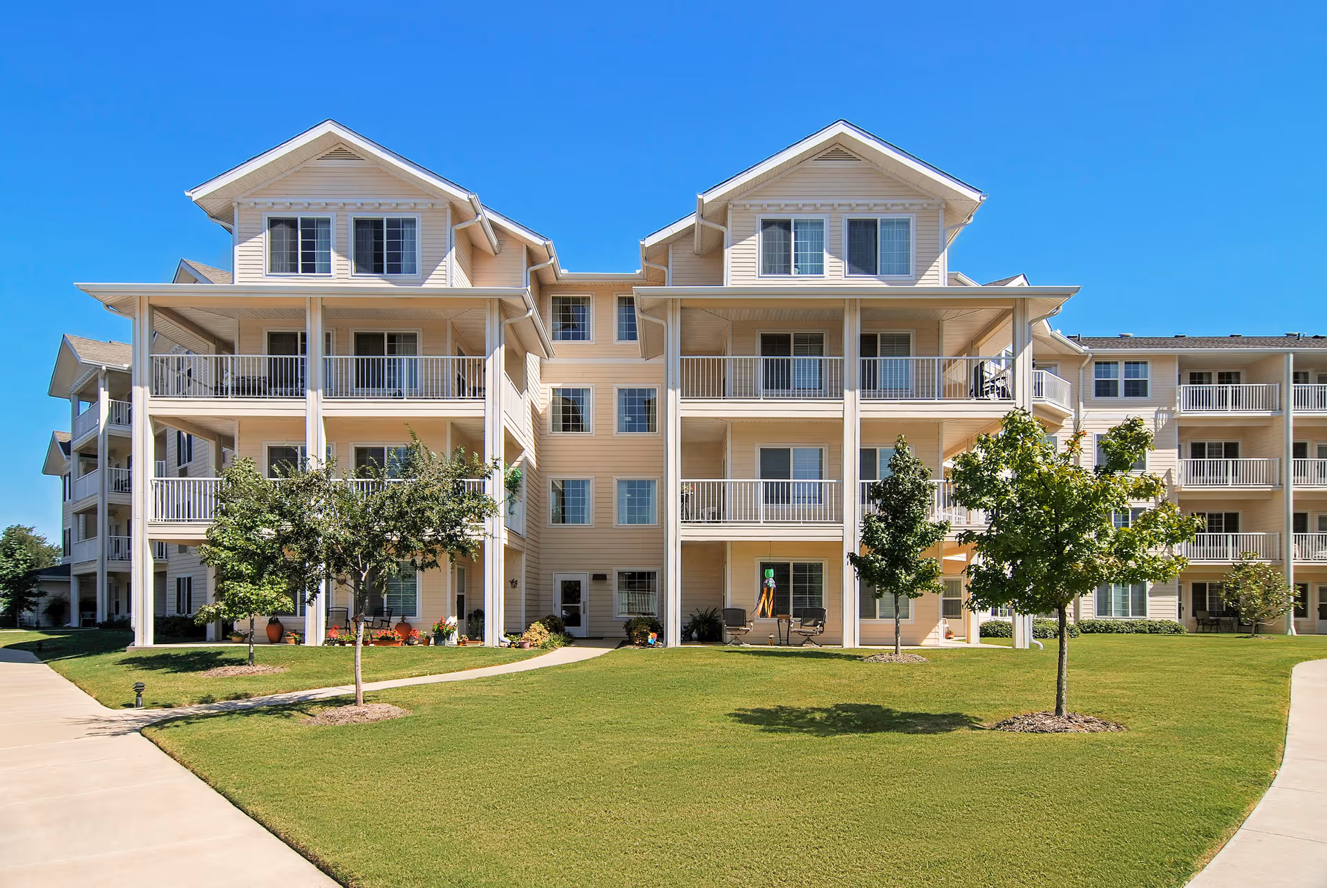 Exterior view of a multi-story senior living facility building with beige siding, multiple balconies, and a well-maintained green lawn with small trees in front under a clear blue sky.