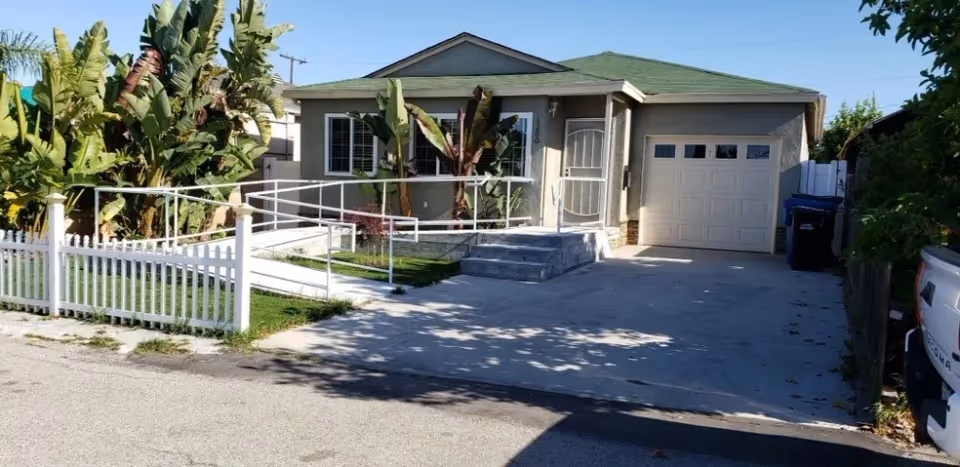 Front exterior of a single-story house with a wheelchair ramp, small porch, garage and driveway bordered by tropical plants and a white picket fence.