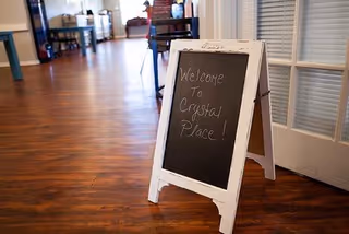 An A-frame chalkboard sign reading "Welcome To Crystal Place!" stands on a wood‑floored hallway inside the facility.