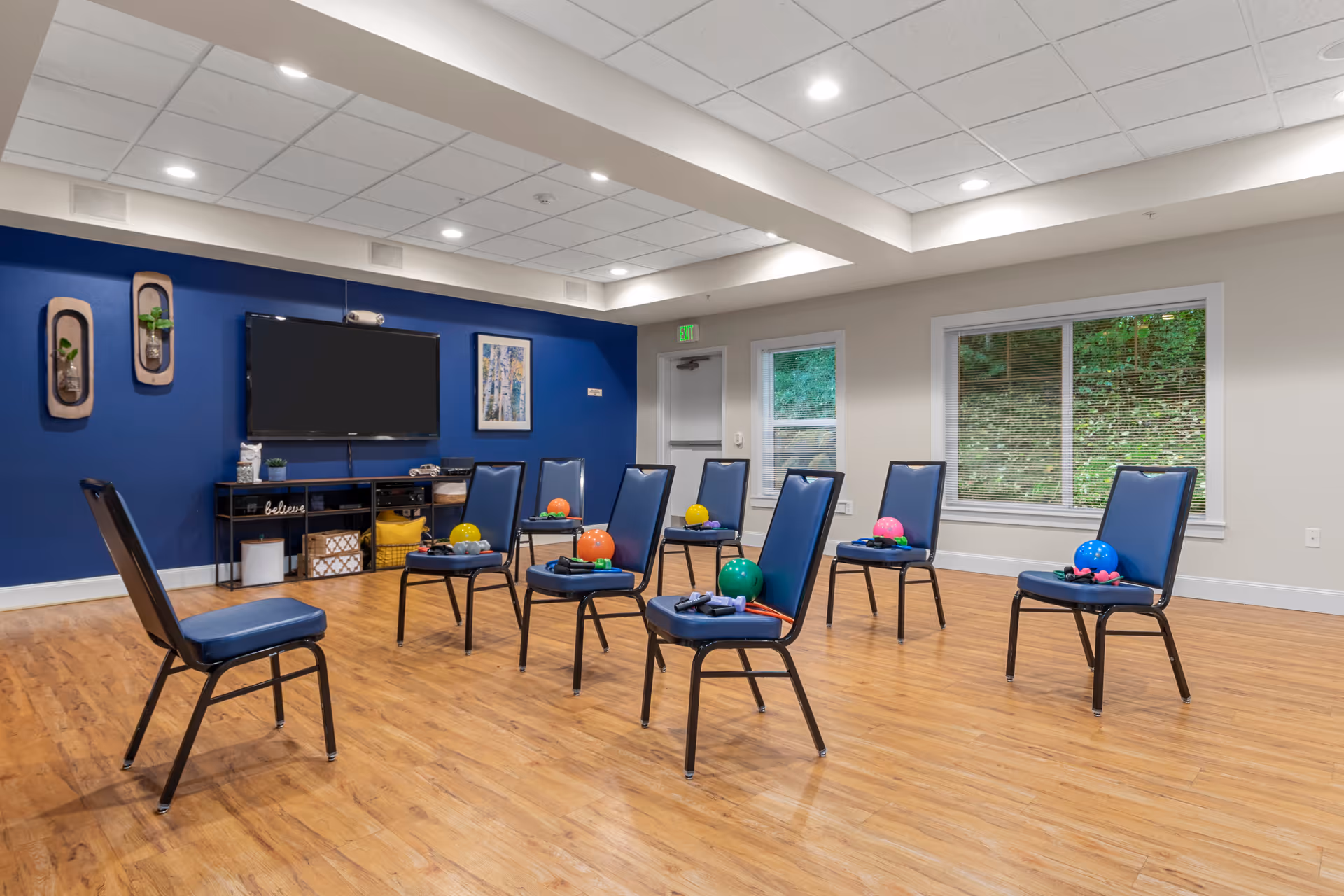 A bright activity room with chairs arranged in a circle, each holding colorful exercise balls and equipment, a TV on a blue accent wall and wood floors.