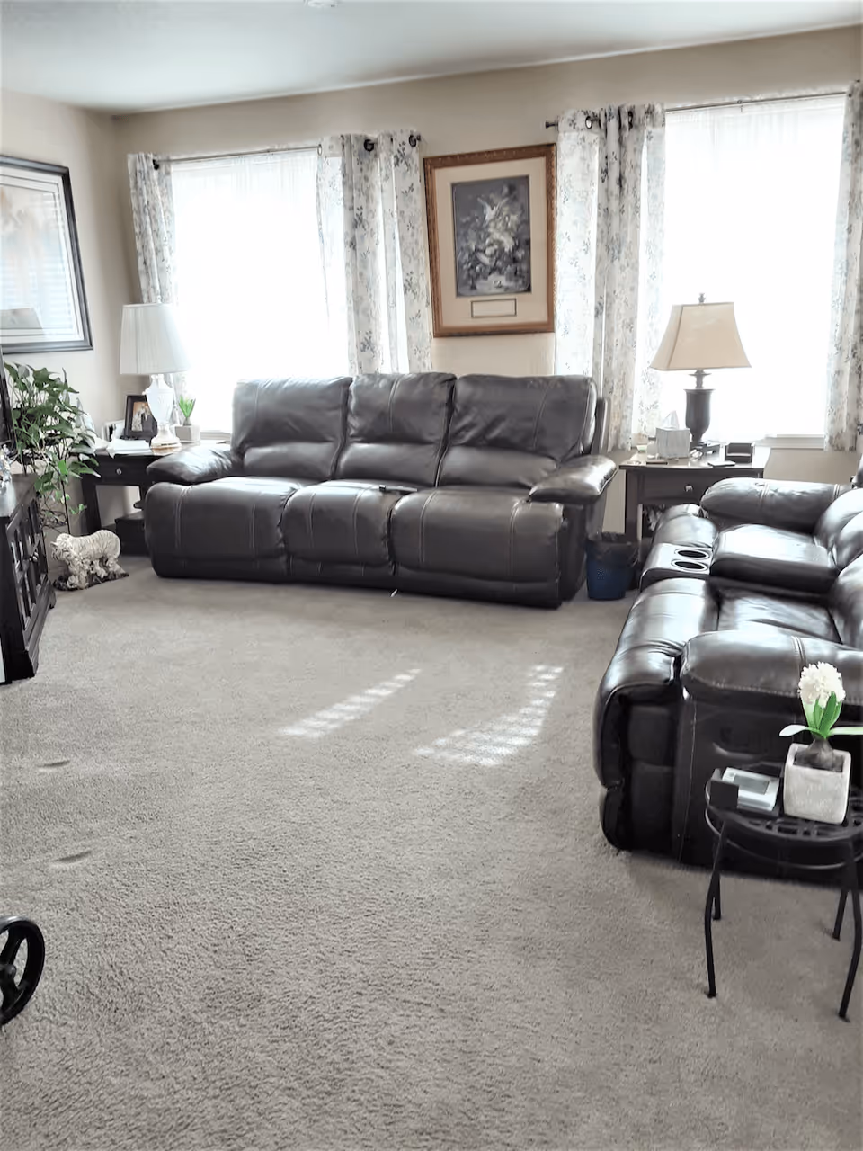 Bright living room with two dark leather sofas, side tables with lamps, framed artwork, and a carpeted floor.