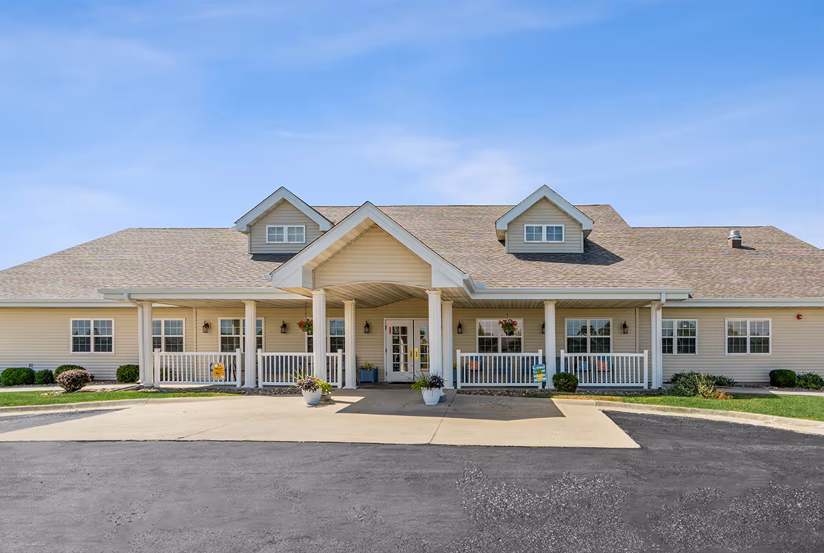 Front exterior view of a single-story senior living facility building with beige siding, a covered entrance supported by white columns, two dormer windows on the roof, and a paved driveway in front. There are potted plants and hanging flower baskets near the entrance.
