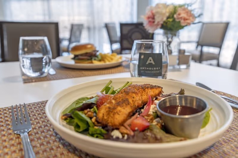Plated salad topped with a piece of grilled fish and dressing on a set dining table with a burger, water glasses, and flowers in a bright dining room.