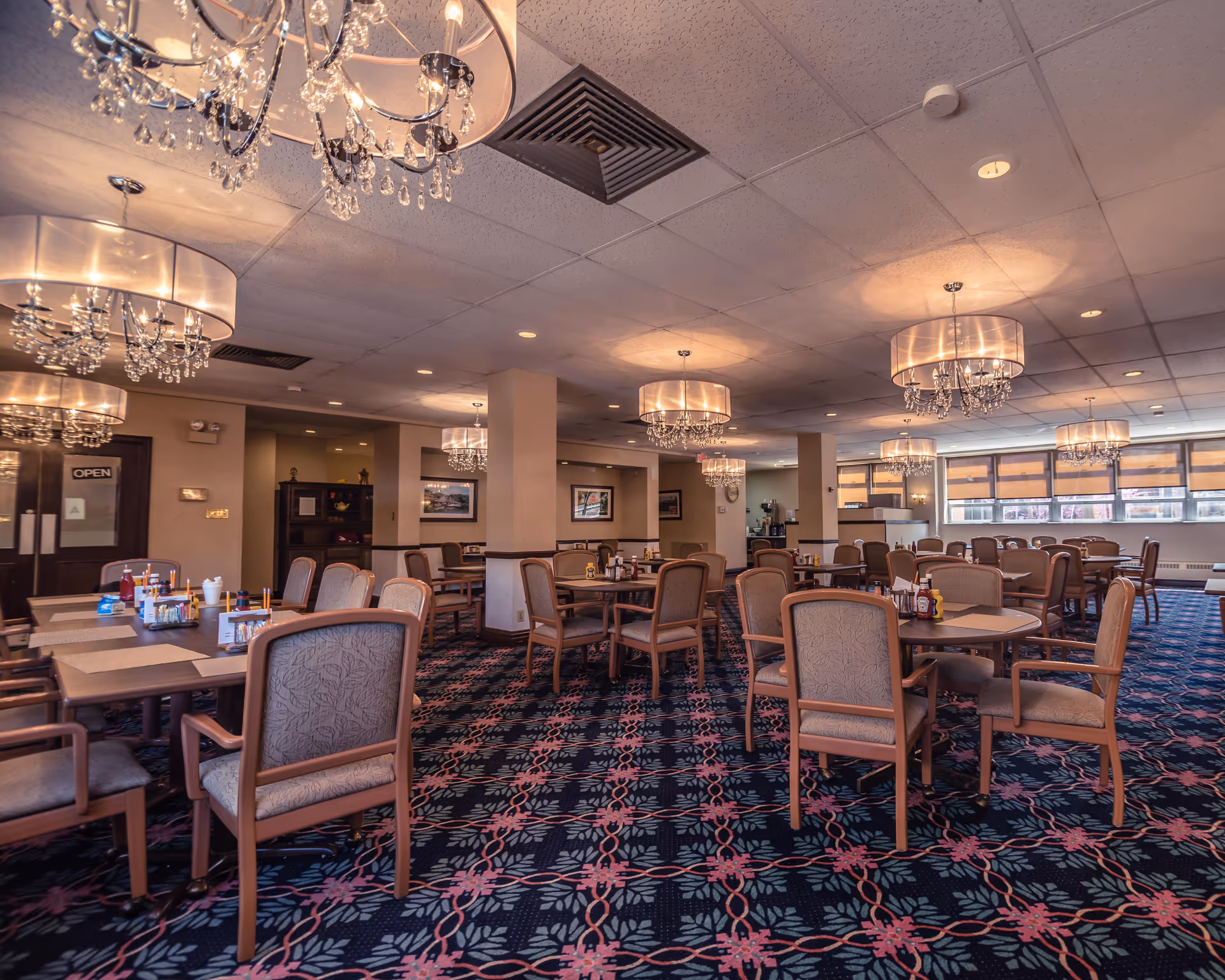 Spacious dining room with multiple tables and chairs arranged neatly. The room features patterned carpet flooring, large windows with blinds, and elegant chandeliers hanging from the ceiling. Tables are set with condiments and napkins, and the room has a warm, inviting atmosphere.