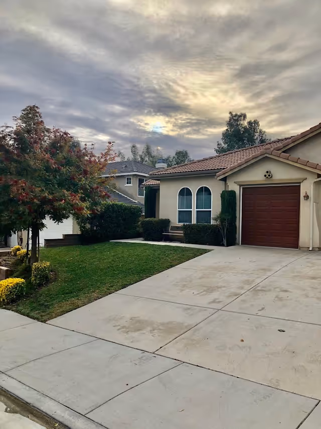 Front exterior of a single-story house with a driveway, garage, lawn, and cloudy sky.