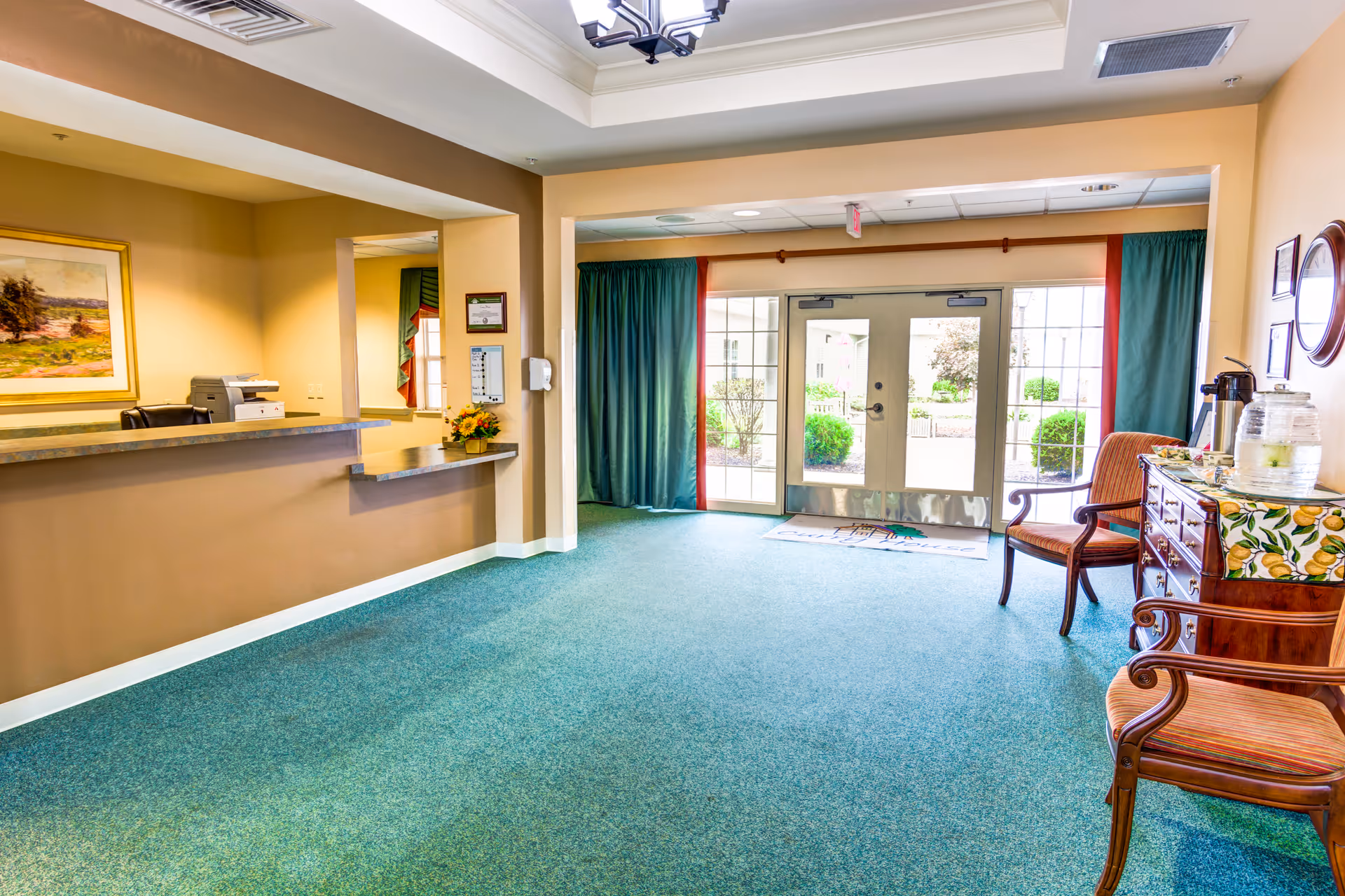 A bright and welcoming reception area with a green carpet, beige walls, and a counter with a printer and flowers. There are two wooden chairs with orange cushions and a wooden sideboard with a lemon-patterned cloth, a water dispenser, and a coffee pot. Large glass doors with green and red curtains lead outside to a garden area.