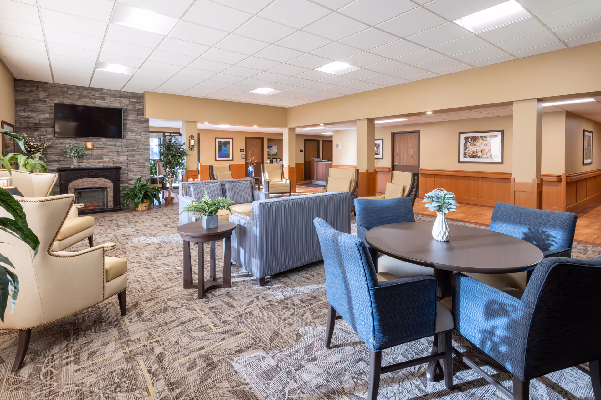 A spacious and well-lit common area in a senior living facility featuring comfortable seating including armchairs, sofas, and a round table with four blue upholstered chairs. The room has a carpeted floor with a patterned design, a stone fireplace with a mounted flat-screen TV above it, and several potted plants. The walls are painted beige with wooden wainscoting, and framed artwork is displayed on the walls. The ceiling has recessed lighting and a grid pattern of ceiling tiles.