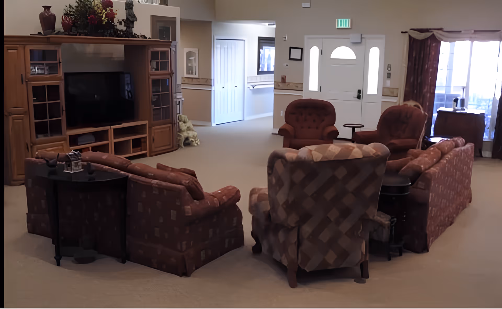 A cozy living room area with multiple upholstered chairs and sofas arranged around a central space. There is a large wooden entertainment center with a flat-screen TV, decorative items on top, and a hallway leading to other rooms. The room has beige carpeting, a white front door with sidelights, and a window with curtains letting in natural light.