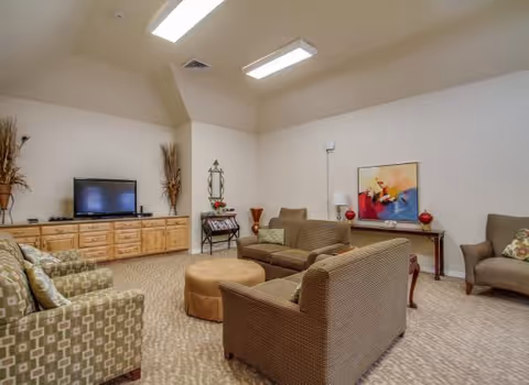 A cozy living room area with beige walls and carpeted floor. The room features a flat-screen TV on a wooden cabinet, two patterned armchairs, a brown sofa, a matching loveseat, and a round ottoman in the center. Decorative items include a colorful abstract painting on a console table, two red vases, a floor lamp, and tall dried plant arrangements in the corners.