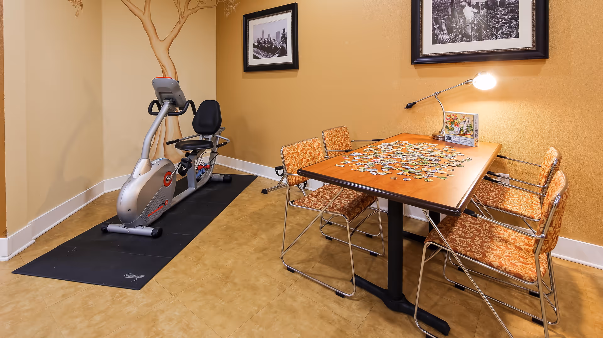 A room with a recumbent exercise bike on a black mat next to a wooden table with four patterned chairs. The table has a partially completed jigsaw puzzle and a lamp on it. The walls are painted yellow with framed black and white photographs hanging.