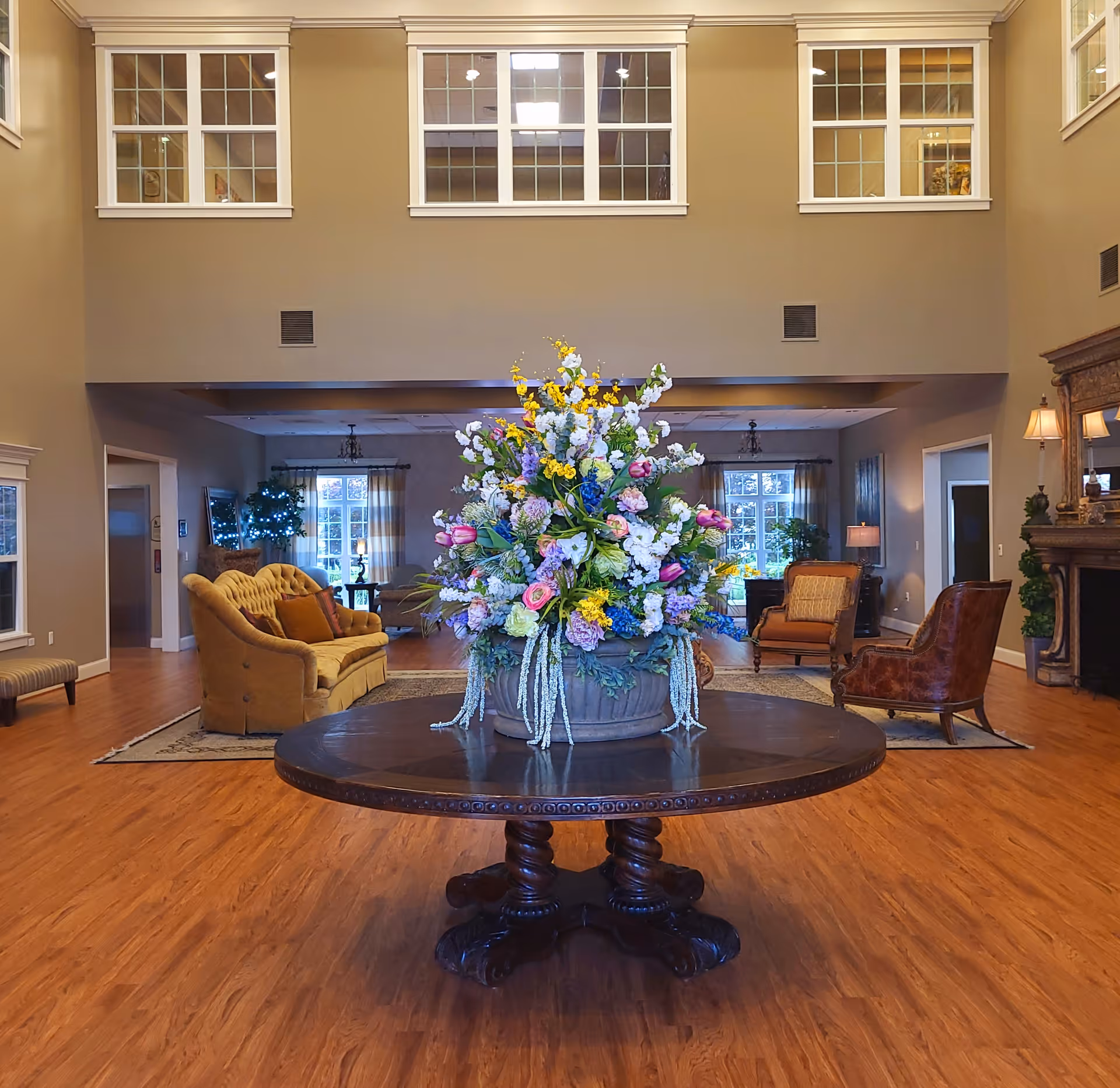 A spacious senior living lobby with a large round table topped by a tall floral arrangement and seating areas and a fireplace in the background.