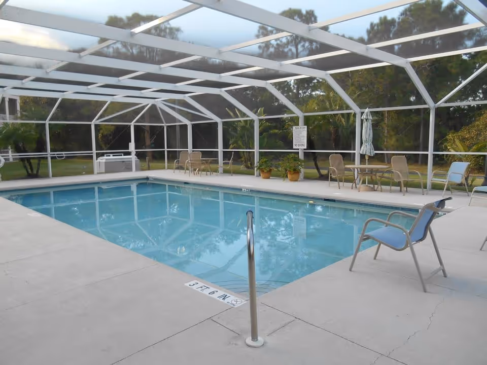 A screened outdoor swimming pool with deck chairs, tables and potted plants around it.