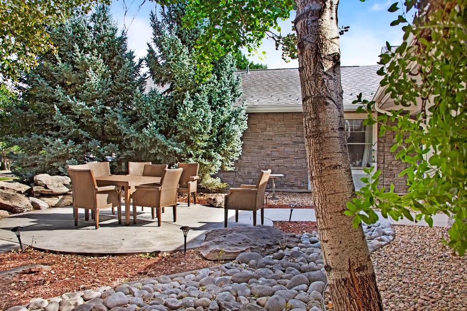 Outdoor patio with a round table and wicker chairs surrounded by trees, rock landscaping, and a stone-faced building wall.