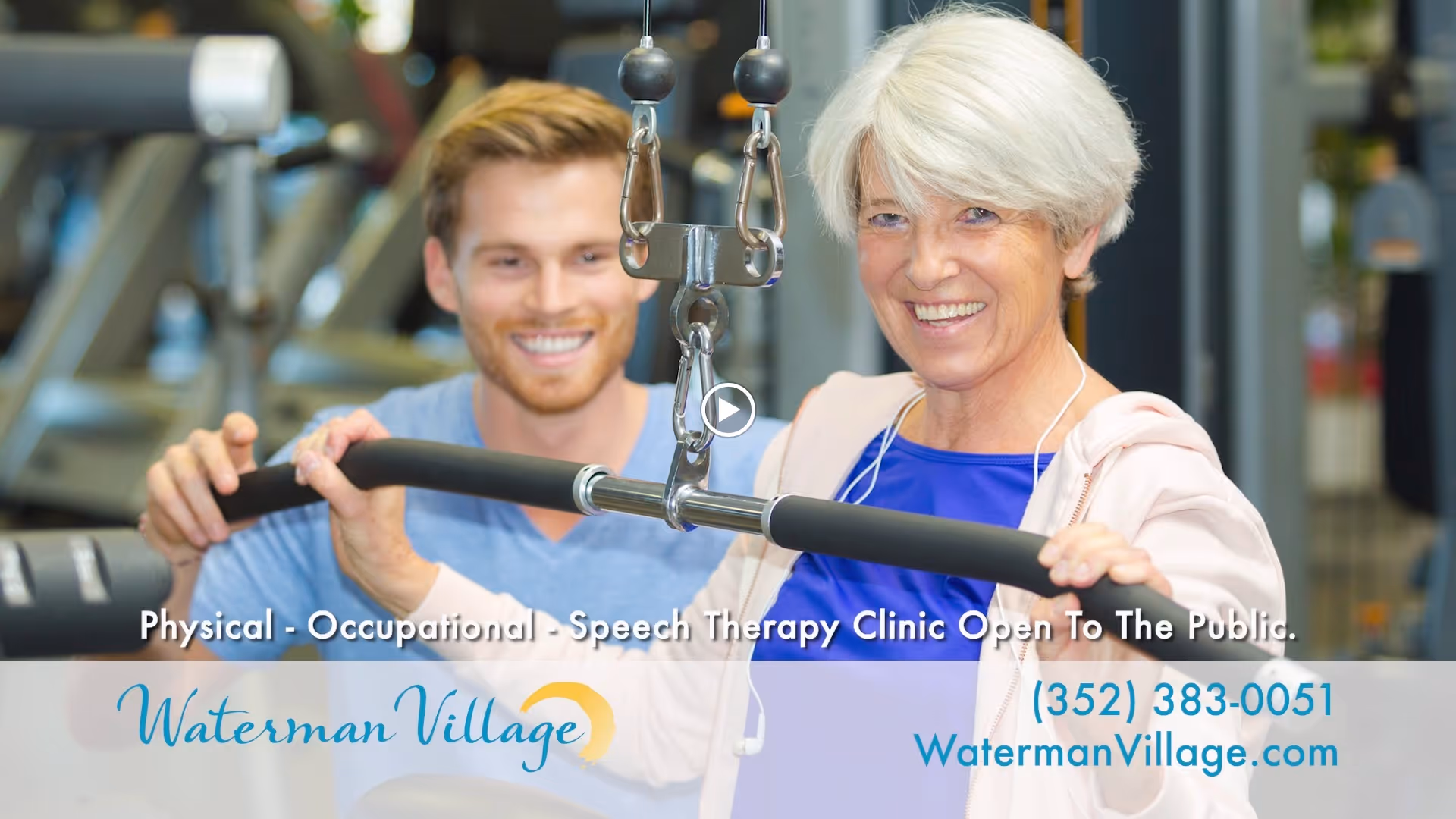 An elderly woman with white hair is smiling while using a lat pulldown machine in a gym, assisted by a younger male trainer who is also smiling. The background shows gym equipment.