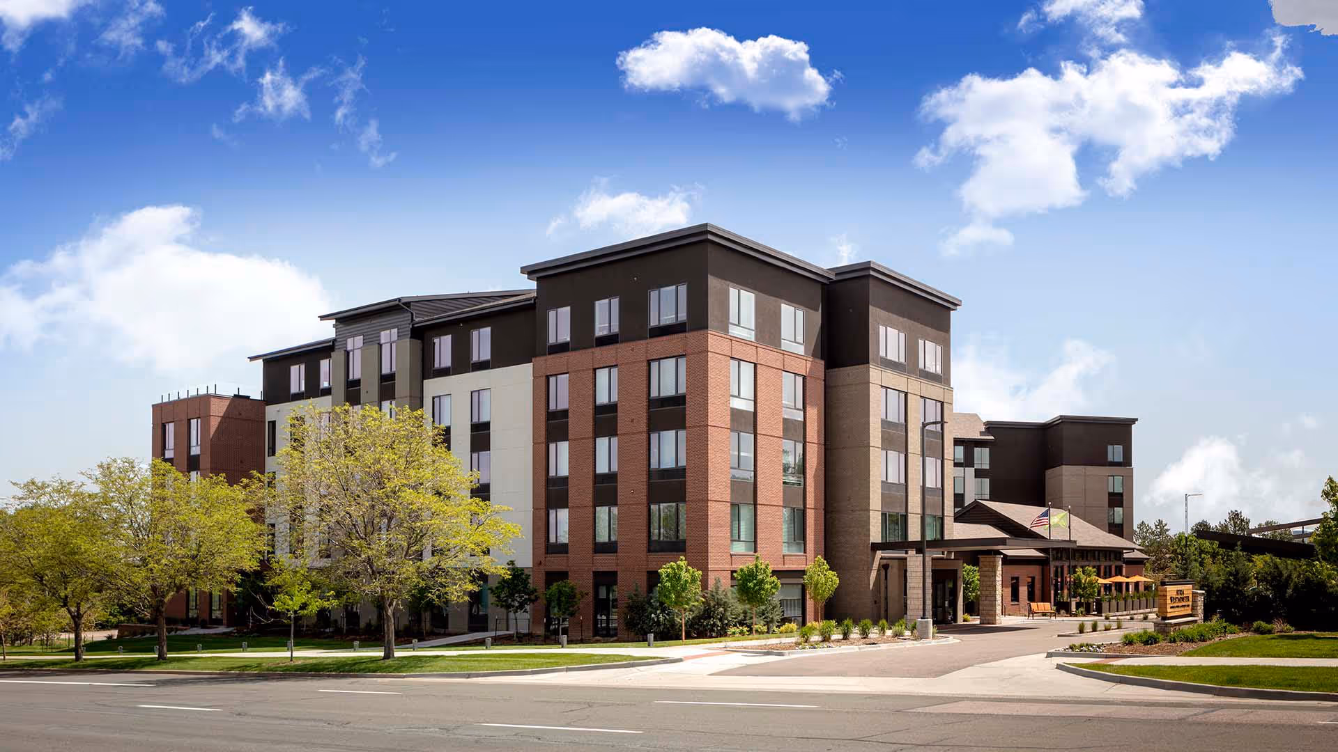 Exterior view of a modern multi-story senior living facility building with a combination of brick and beige siding, surrounded by trees and landscaping under a partly cloudy blue sky.
