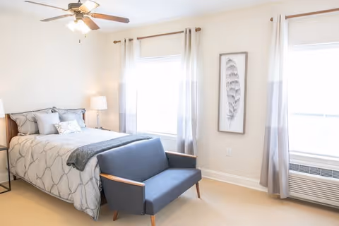A bright bedroom with a bed dressed in gray and white bedding, a small blue loveseat at the foot of the bed, two large windows with white and gray curtains, a ceiling fan with lights, a floor lamp, and a framed feather artwork on the wall.