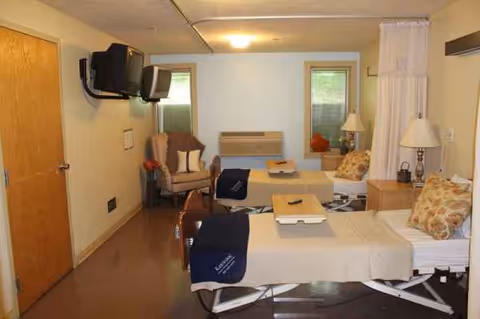Two hospital beds in a shared patient room with chairs, lamps, and small windows.