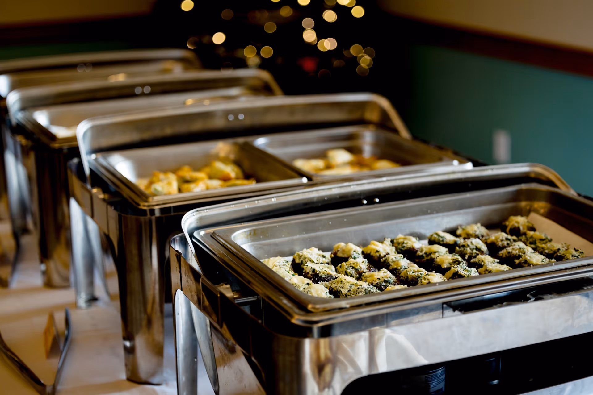 A buffet setup with multiple stainless steel chafing dishes filled with various cooked foods, arranged in a row on a table with a blurred background featuring warm lighting.