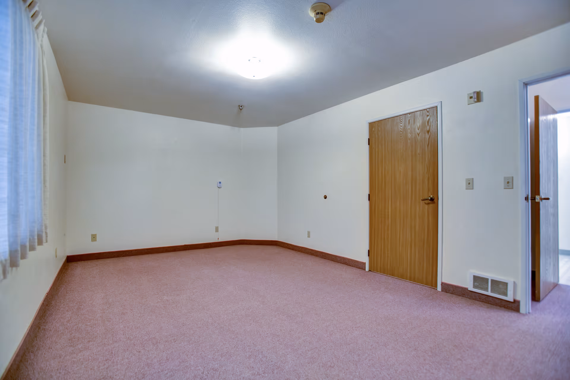 Empty room with light pink carpet, white walls, a wooden door, and a window with sheer curtains on the left side. The ceiling has a round light fixture and a smoke detector.