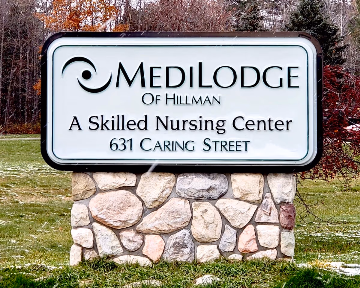 A stone-based roadside sign that reads "MediLodge of Hillman — A Skilled Nursing Center — 631 Caring Street" on a grassy lawn.