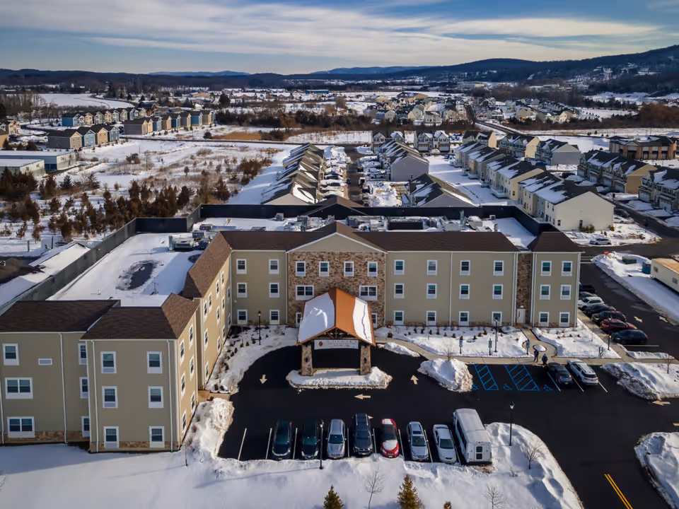 Aerial view of a three-story senior living building with a covered entrance, snow-covered parking lot, and surrounding residential neighborhood.