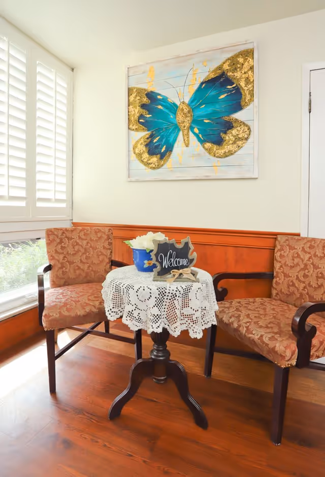 Two patterned chairs flank a lace-draped table beneath a butterfly painting in a bright waiting area.