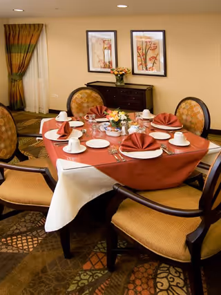A dining table set for four with a red tablecloth and white undercloth, each place setting includes a folded red napkin, white plates, cups, and silverware. The table is surrounded by four cushioned chairs with patterned upholstery. In the background, there is a sideboard with a flower arrangement and two framed floral artworks on the wall.