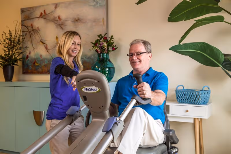 A man using a NuStep exercise machine with the assistance of a female caregiver or therapist in a well-decorated room with plants and artwork on the wall.