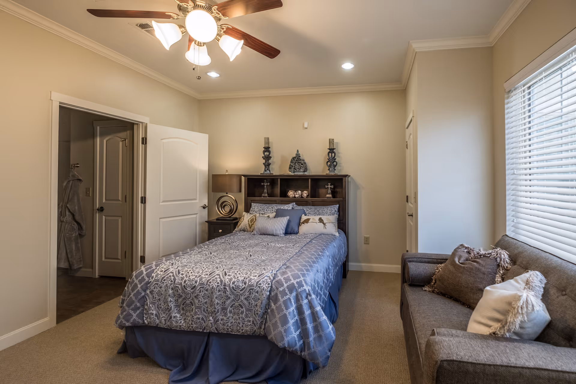 A cozy bedroom with a bed covered in a blue and white patterned comforter and multiple pillows. There is a dark wooden headboard with decorative items on top, a nightstand with a lamp, a ceiling fan with lights, a gray sofa with cushions near a window with blinds, and an open door leading to another room.