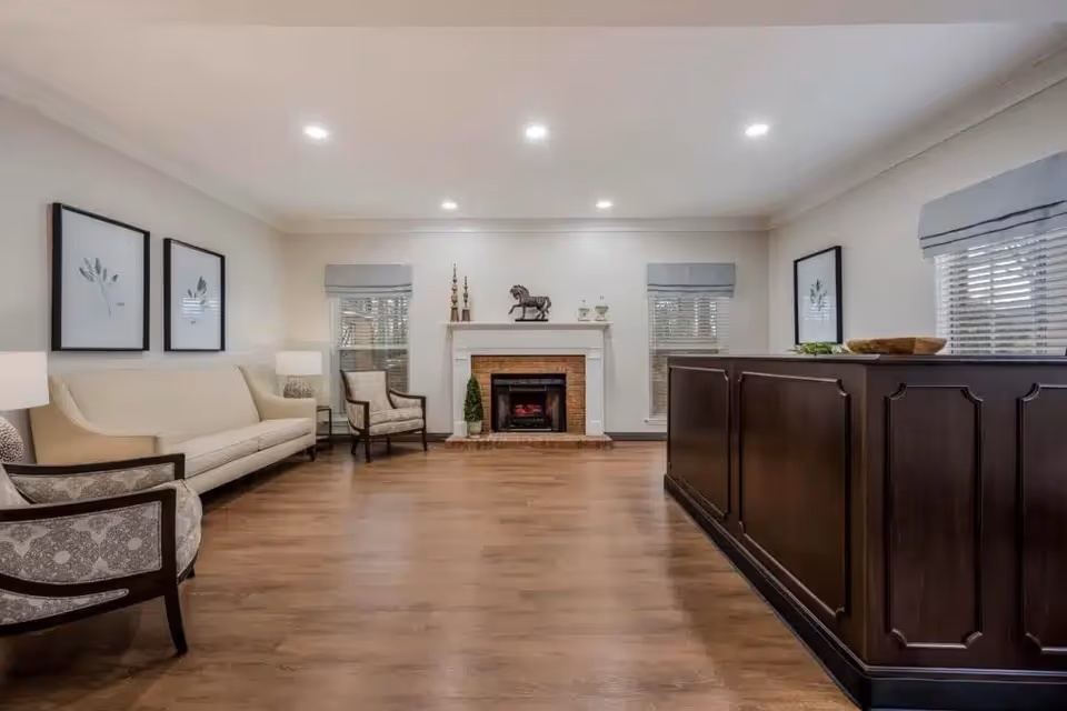 A spacious and well-lit living room with wooden flooring, a beige sofa, two patterned armchairs, a dark wooden reception desk or counter on the right, and a white fireplace with decorative items including a horse statue and candlesticks. The room has three windows with blinds and light gray valances, and two framed botanical prints on the walls.