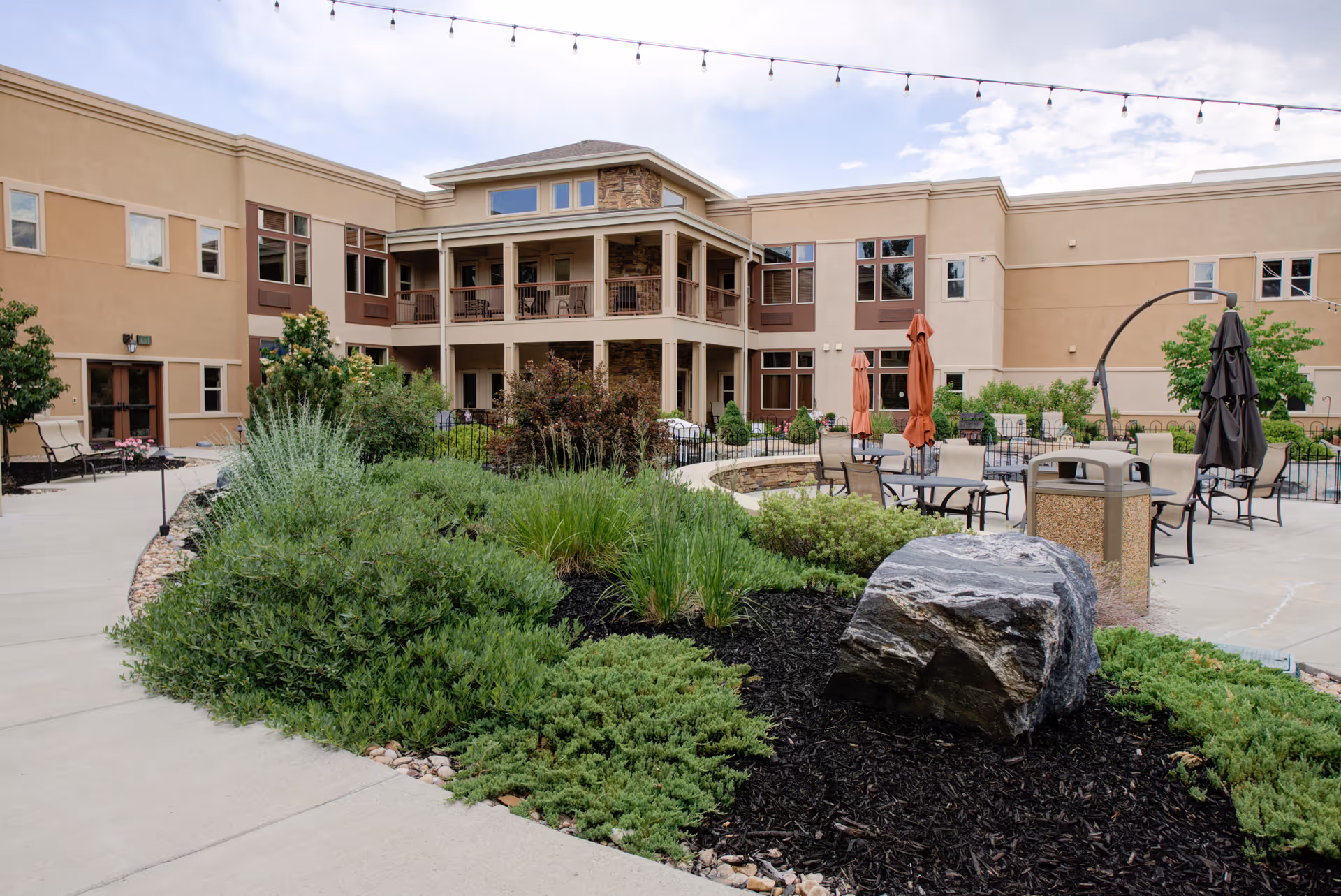 Outdoor courtyard area of a senior living facility with landscaped greenery, a large rock, patio tables with umbrellas, and a two-story building with balconies in the background under a partly cloudy sky.