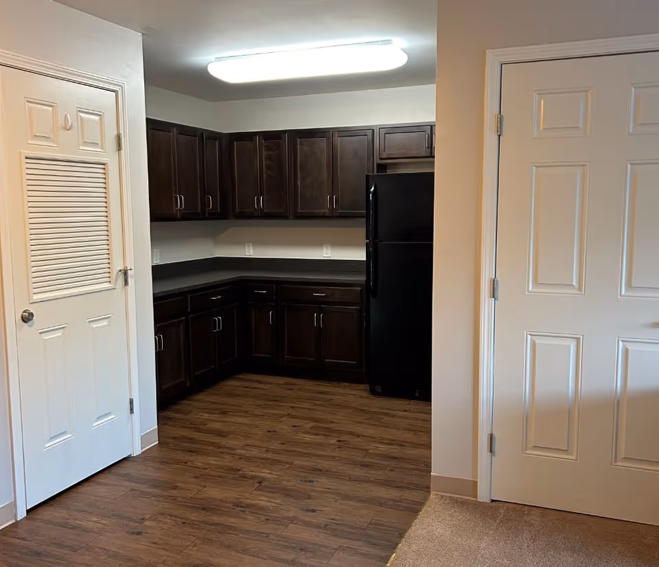 Interior view of a kitchen area with dark wooden cabinets, black refrigerator, and wood flooring. There are two white doors visible, one on the left with a vent panel and one on the right. The walls are light-colored and the ceiling has a rectangular fluorescent light fixture.