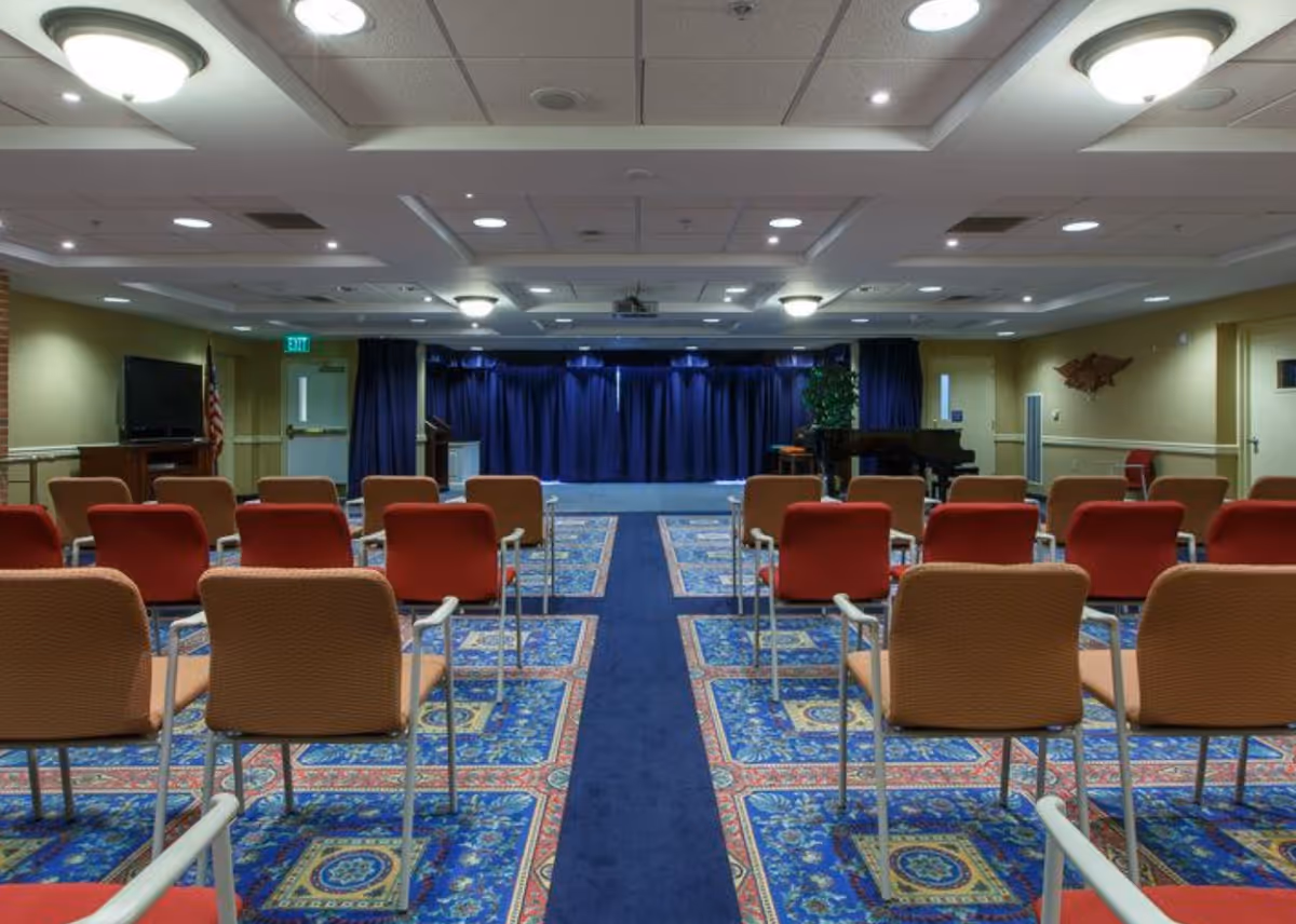 A conference or meeting room with rows of chairs facing a stage with blue curtains. The room has a patterned blue carpet with red and beige chairs, ceiling lights, a piano on the right side, and an American flag near a television on the left side.