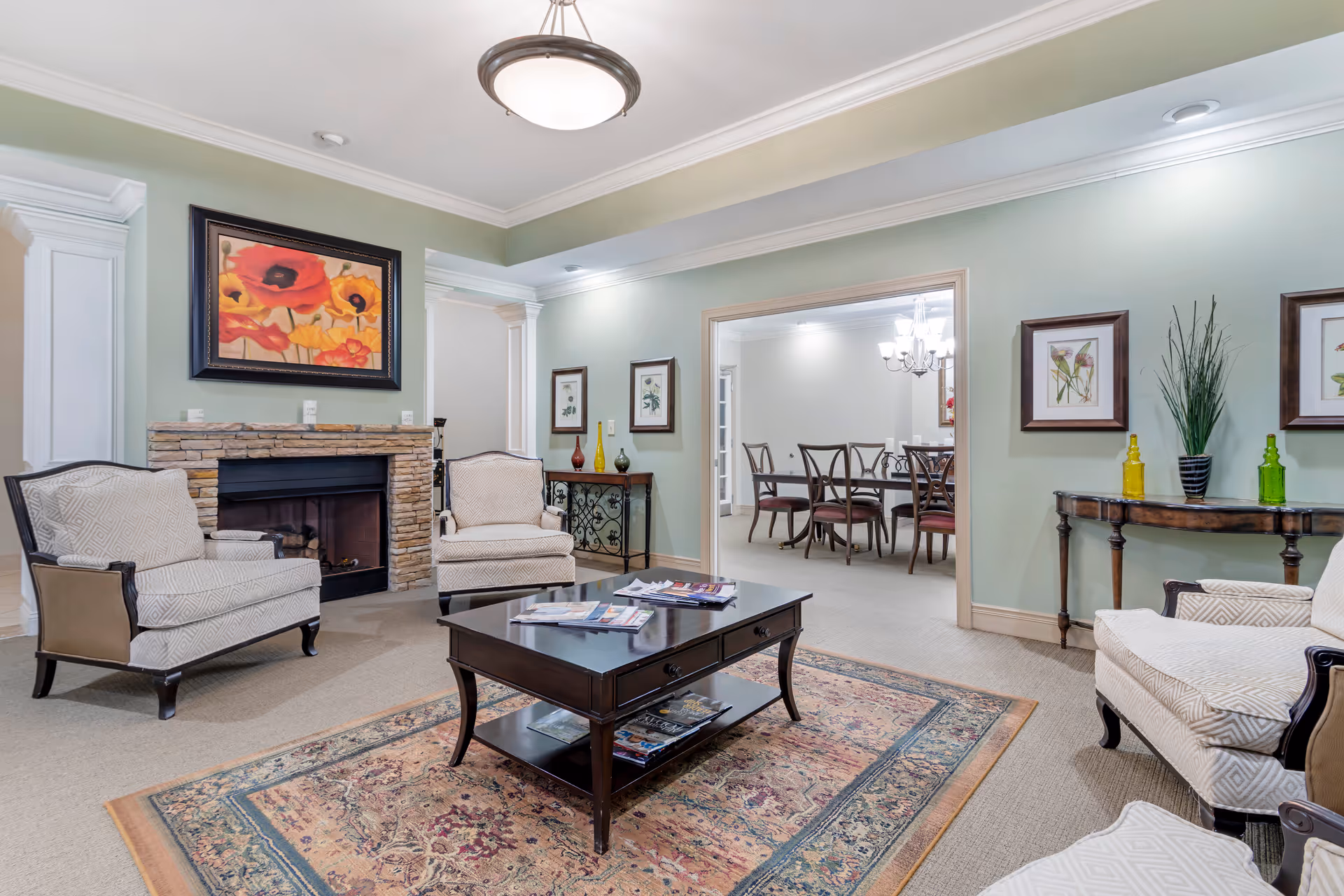 A cozy living room area with three upholstered armchairs arranged around a dark wooden coffee table on a patterned rug. The room features a stone fireplace with a colorful floral painting above it. The walls are painted light green and decorated with framed botanical prints. A doorway leads to a dining room with a table and chairs under a chandelier. The room has soft lighting and elegant decor.