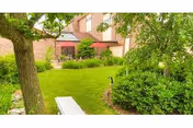 Landscaped courtyard with a grassy lawn, trees and shrubs, a bench in the foreground and a patio with red umbrellas by a brick building.