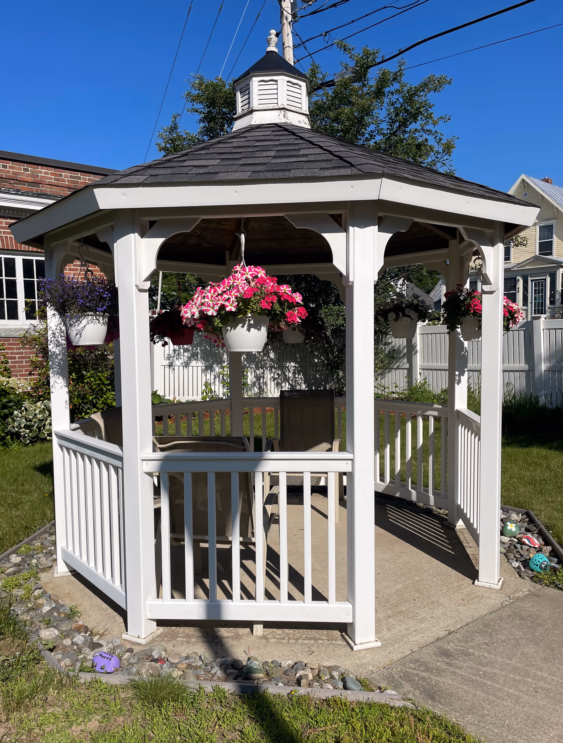 White wooden gazebo with hanging pink and purple flower baskets and patio chairs on a sunny lawn near a white fence and brick building.