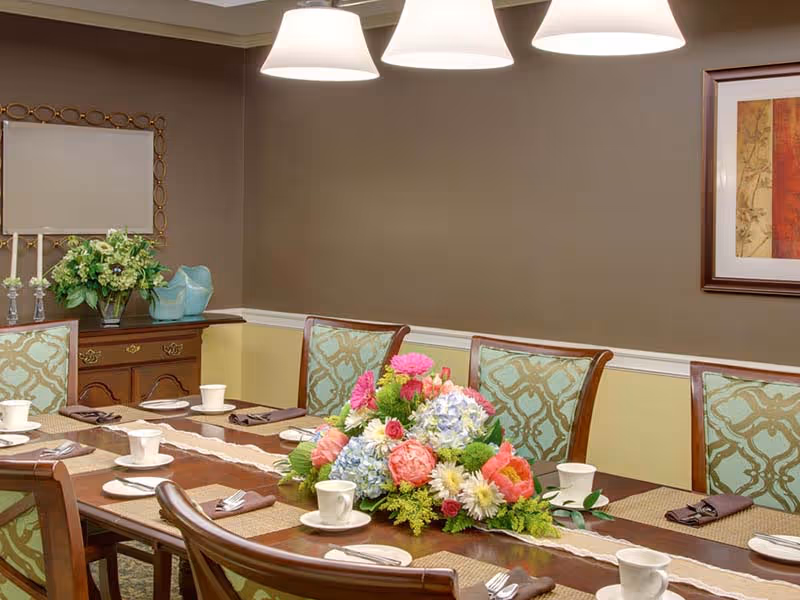 A dining room with a long wooden table set with white cups, saucers, and silverware wrapped in brown napkins. The table has a centerpiece of colorful flowers including pink, blue, and white blooms. The room features upholstered chairs with a blue and beige pattern, a sideboard with a green floral arrangement, and framed artwork on the wall. Three pendant lights hang from the ceiling above the table.