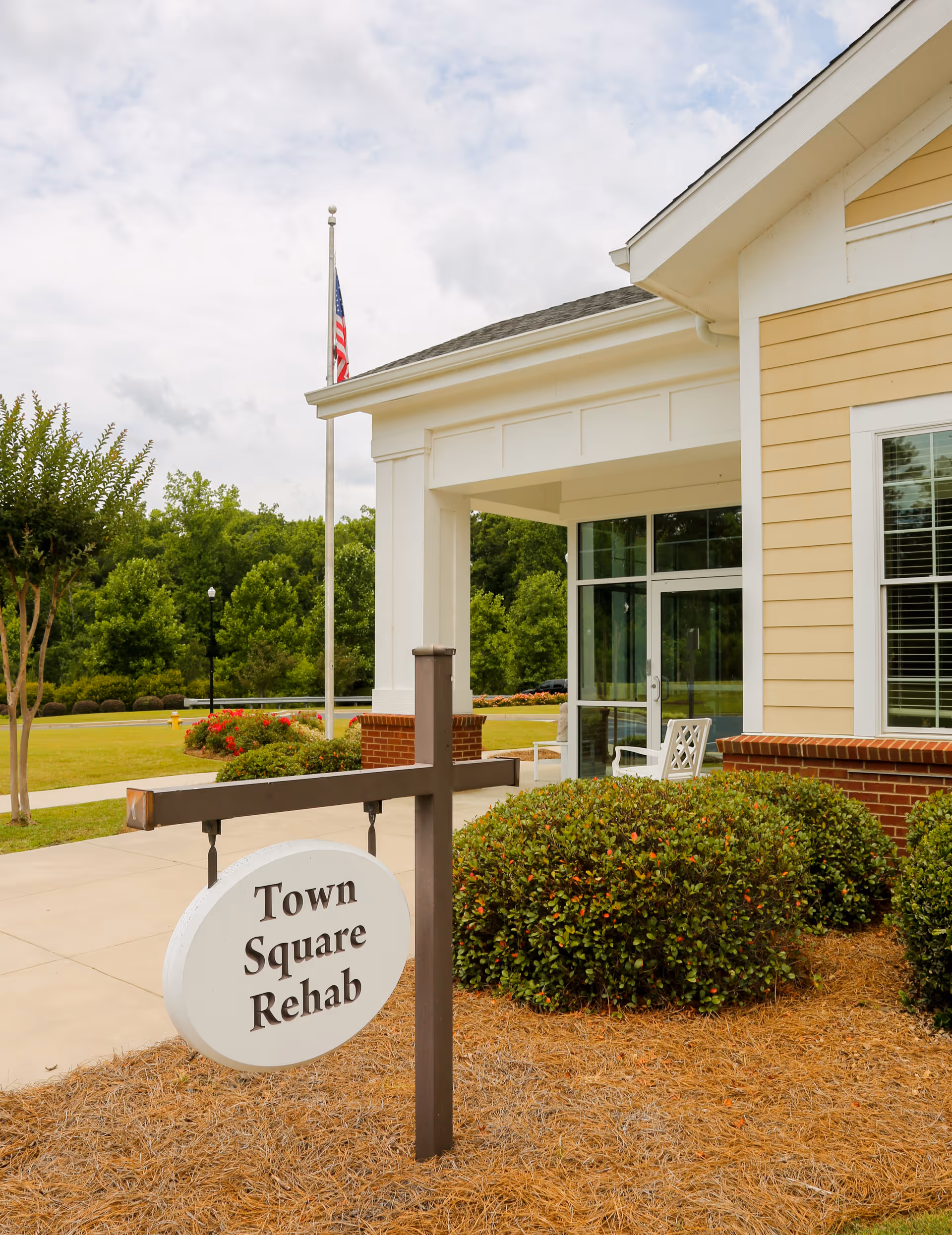Exterior view of a rehabilitation facility with a sign that reads 'Town Square Rehab' in front of a building with yellow siding, white trim, and large windows. There are bushes and a flagpole with an American flag near the entrance.