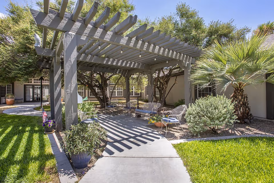 Outdoor seating area at Brookdale East Tucson featuring a gray wooden pergola casting shadows on a concrete pathway. The area includes metal chairs with blue cushions and a bench, surrounded by green grass, potted plants, trees, and desert landscaping under a clear blue sky.