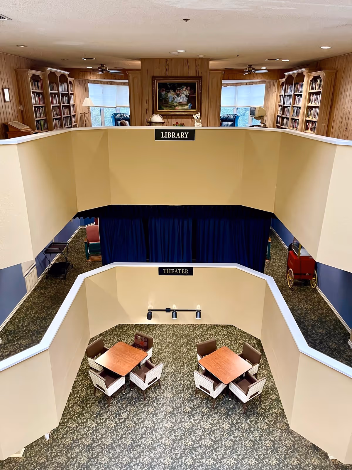 Interior view of a senior living facility showing a two-level space with a library on the upper level featuring bookshelves, armchairs, and a painting on the wall. The lower level is labeled as a theater area with two square tables, each surrounded by four chairs, and a patterned carpet covering the floor.