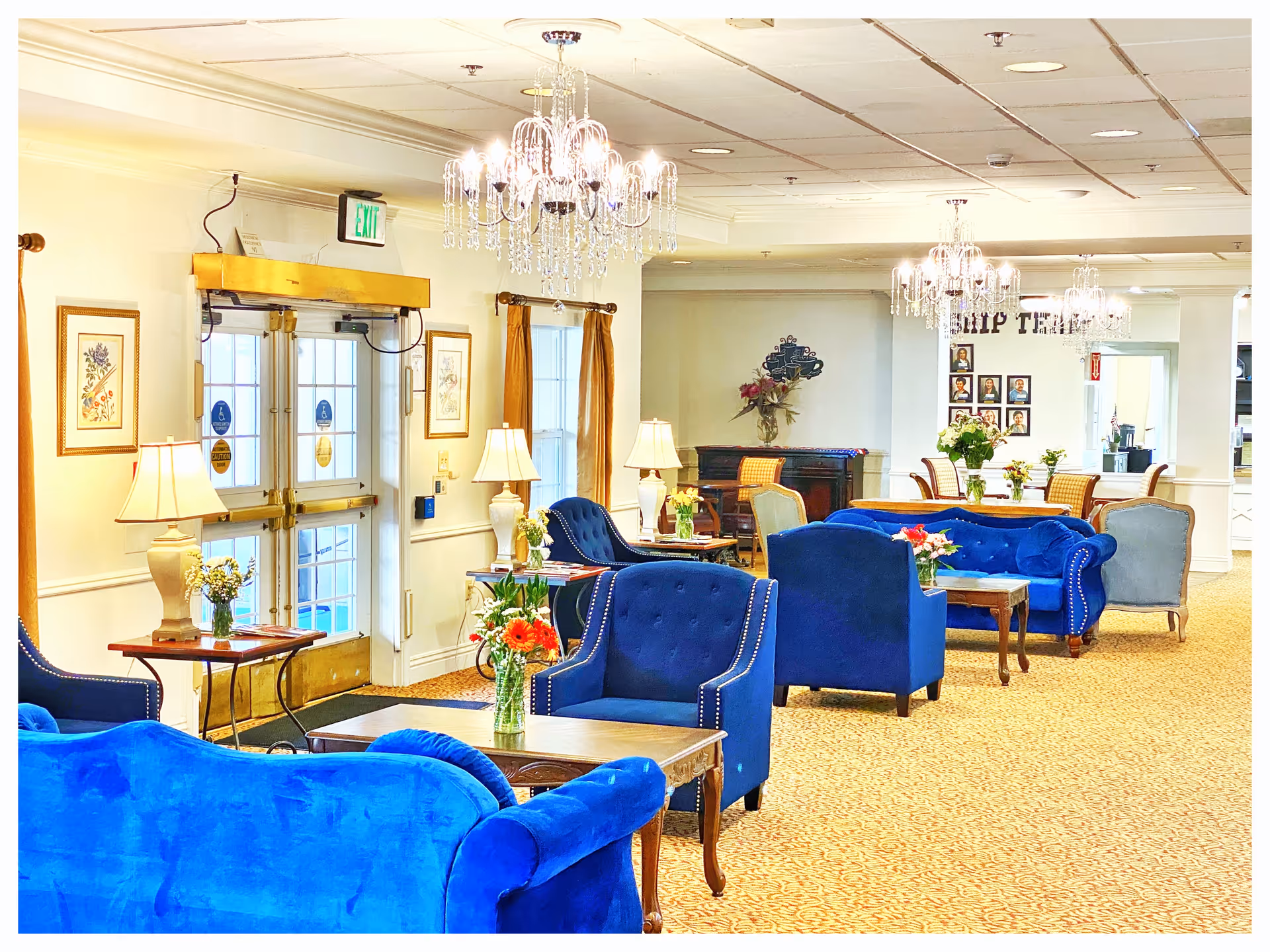 Bright common lounge with blue velvet sofas and armchairs, chandeliers, side tables, and floral arrangements.