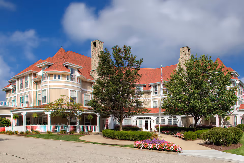 Exterior view of a large senior living facility building with beige siding and a red roof, surrounded by trees, bushes, and colorful flower beds under a partly cloudy blue sky.
