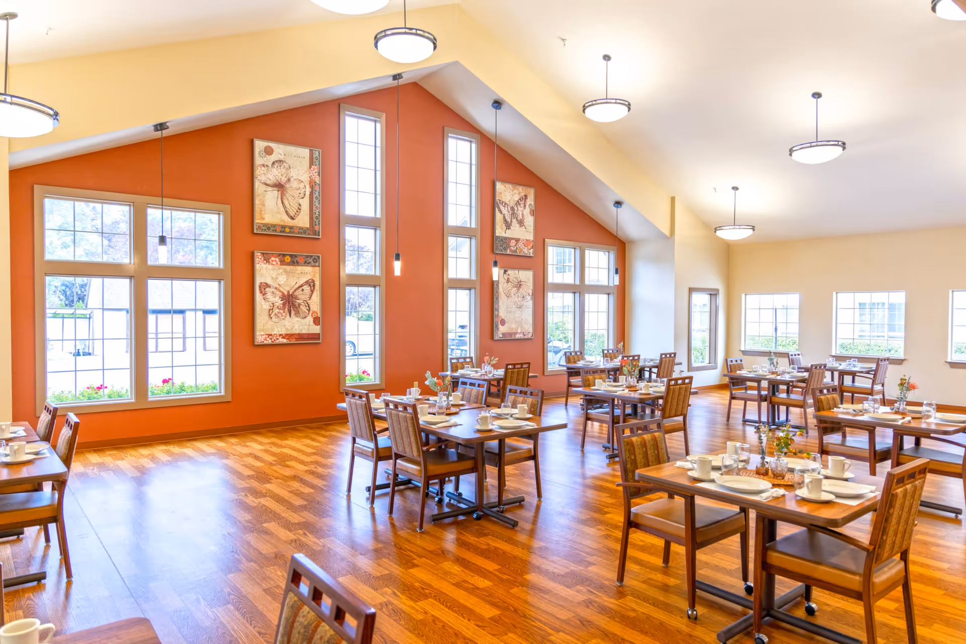 Bright and spacious dining room with wooden floors and multiple tables set with white plates, cups, and silverware. Large windows allow natural light to fill the room, and the walls feature butterfly artwork. Ceiling lights hang from the high, sloped ceiling.