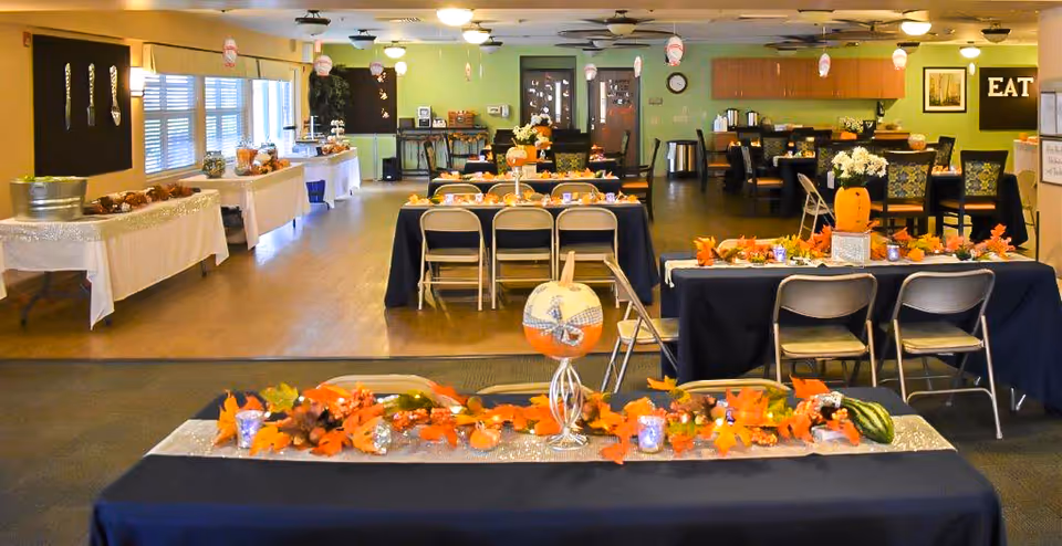 A decorated dining room in a senior living facility with tables covered in dark blue tablecloths and autumn-themed centerpieces including pumpkins and fall leaves. The room has multiple tables and chairs arranged for a gathering or meal, with a buffet table along the left wall. The walls are painted green and there are framed pictures and a sign that says 'EAT' on the right side.