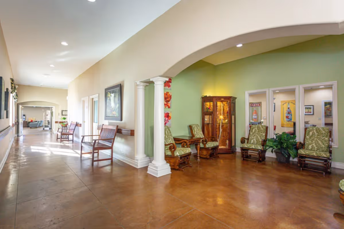 A spacious hallway in a senior living facility with polished brown floors and cream-colored walls. The hallway features wooden benches along the left side and a seating area on the right with four cushioned chairs and a wooden cabinet against a light green wall. Decorative columns and framed artwork are also visible, with natural light streaming in from the far end.