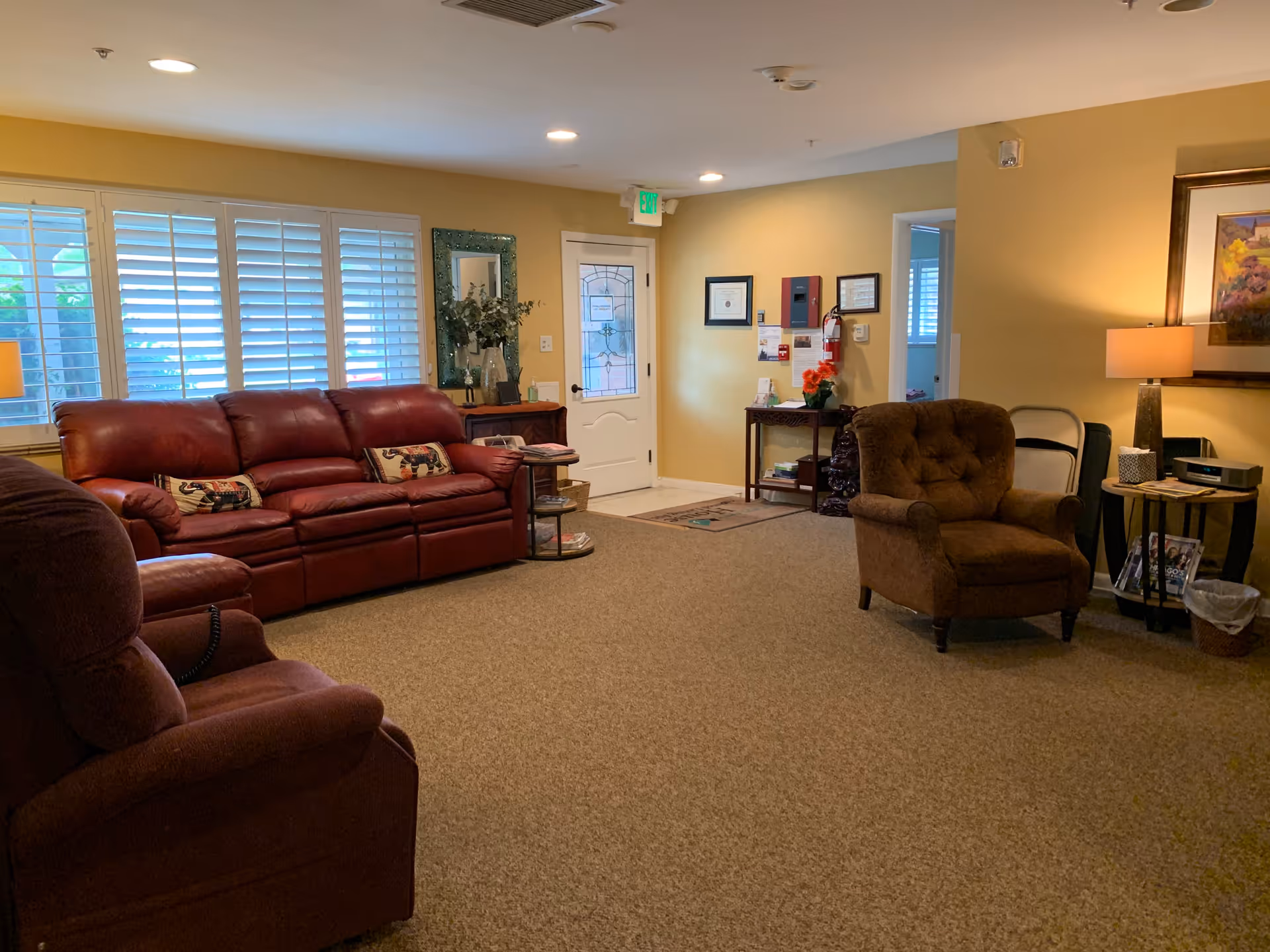 A cozy living room with a brown carpet, a red leather sofa with decorative pillows, a brown armchair, and a side table with a lamp and framed picture on the wall. There is a door with a glass panel and a window with white shutters letting in natural light. A small table with flowers and framed certificates is against the wall near the door.