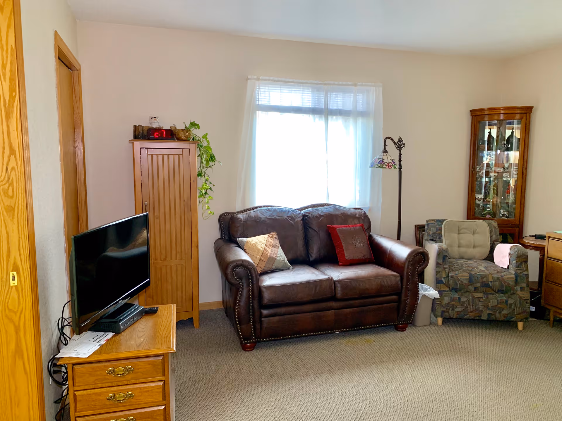 A cozy living room with a brown leather loveseat adorned with two pillows, a patterned armchair with a cushion, a wooden TV stand holding a flat-screen television, a tall wooden cabinet with a plant on top, a floor lamp with a stained glass shade, and a glass display cabinet in the corner. The room has beige walls and carpeted flooring, with a window covered by sheer white curtains letting in natural light.
